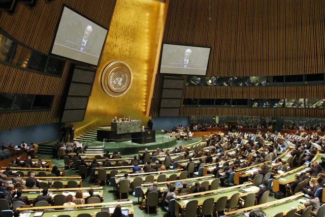 UN assembly hall with people seated watching a speaker on screen