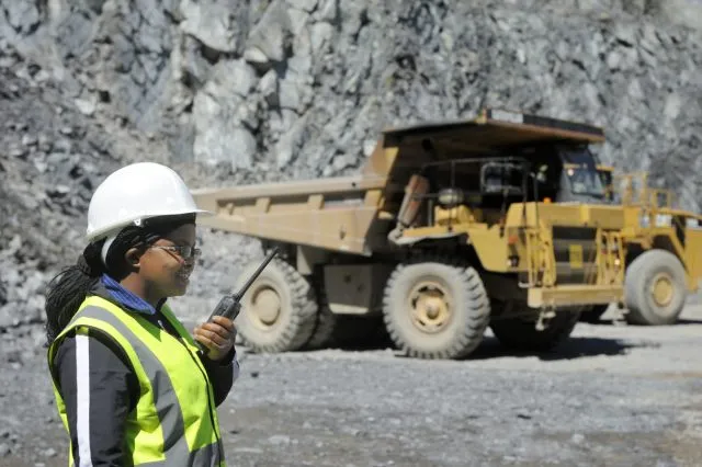 Female mine worker wearing a hi viz jacket and white hard hat talking into a radio next to vehicles