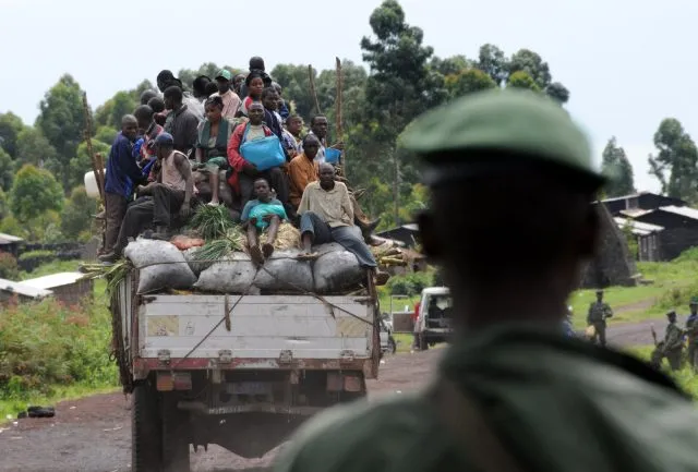People on the back of a vehicle driving away from a soldier who is watching