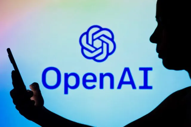 A woman is seen in silhouette holding a phone in front of a sign with a logo for OpenAI, an artificial intelligence company.