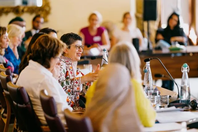 A female speaker, sitting at the conference table addressing a Wilton Park conference
