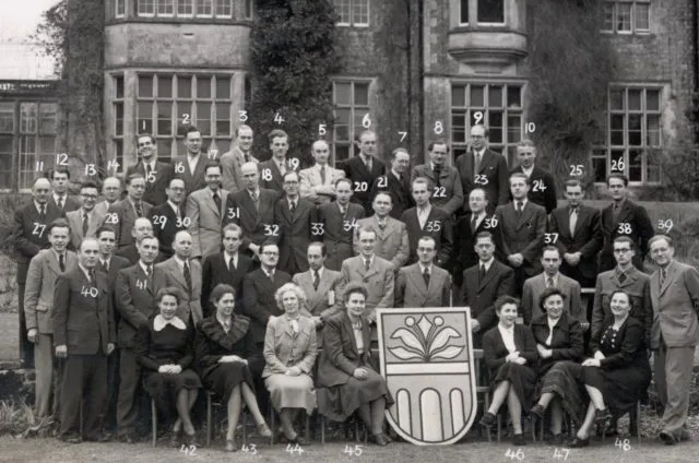 Attendees at an early Wilton Park event are seated for a group photo, with a shield decorated in the Wilton Park logo.