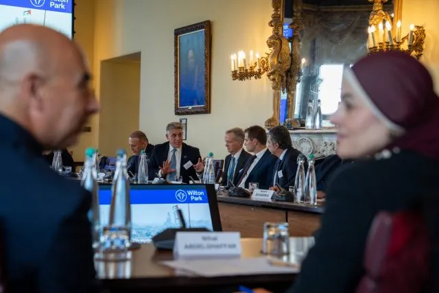 Two participants smile at one another in the foreground, with speakers visible in the back