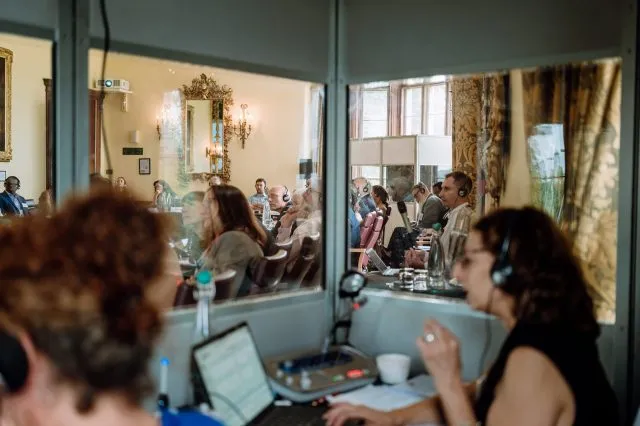The view through a translation booth looking into a conference as participants sit around a large table.