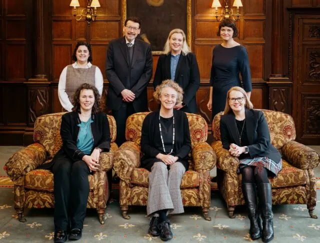 A portrait of the partners for the event, three seated in armchairs and four standing behind the chairs, against a wood panelled background