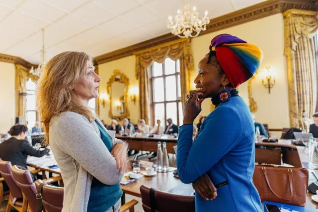 Two female participants talk in the foreground of the conference room, with the table in the background