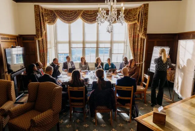 Participants sit a large table in the study at Wilton Park. A participant stands at a board writing notes.