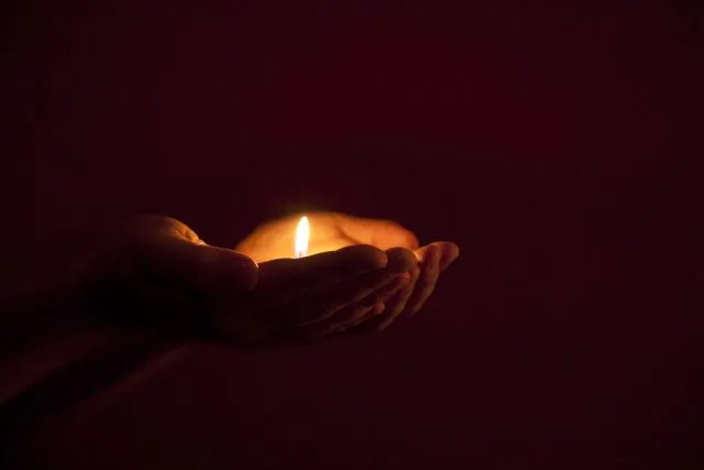 A pair of hands cradle a small candle against a dark backdrop.