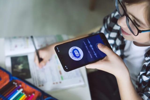 A young person holds a phone in one hand while writing on a document on their desk