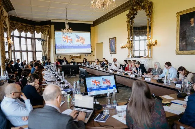 Participants seated around the conference table. Screens showing the UK and US flags are on display in the room