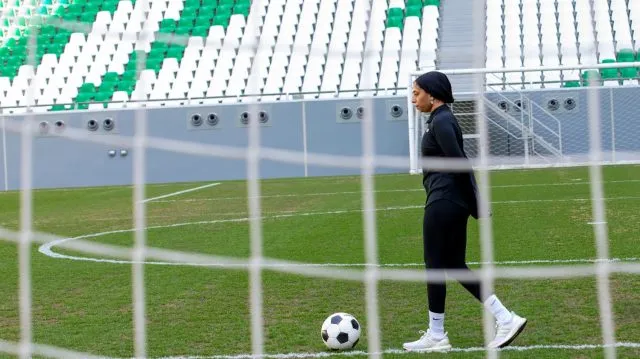 A young woman wearing a hijab and black sportswear walks up to a football on a green football pitch. The photo is taken through football goal netting
