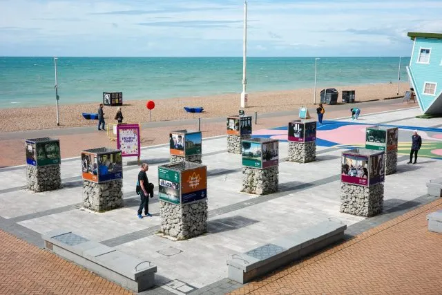 A wide shot of a seafront exhibition in Brighton. Large gabions have images attached on 4 sides and the tall columns stand in a square next to the beach. People wander through and look at the images.
