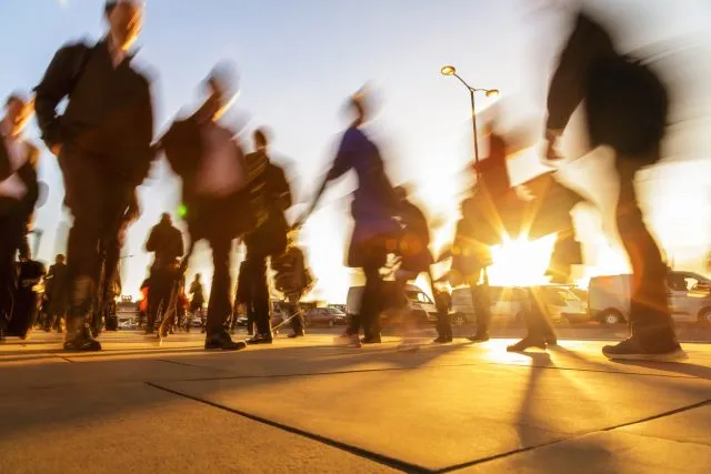 A blurred street scene of people walking across a bridge with bright sunlight behind them.