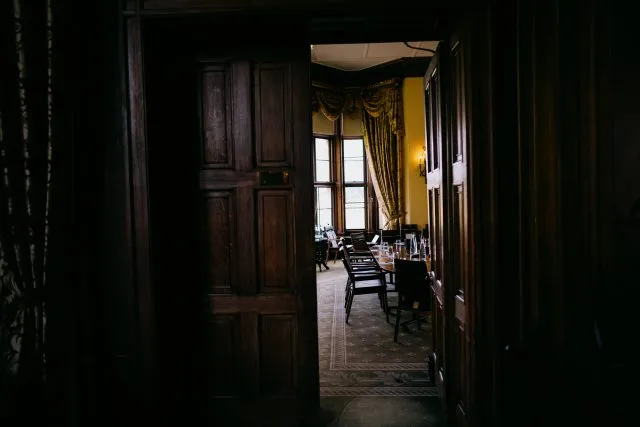 View through a partially open wooden doorway into the conference room at Wilton Park, showing a long meeting table with chairs, water bottles and glasses set out, tall windows letting in daylight, and ornate gold-patterned curtains against warm yellow walls.