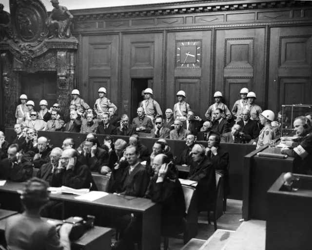 Defendants in the Nuremburg Trials of Major War Criminals are seated in the courtroom, with soldiers standing behind them