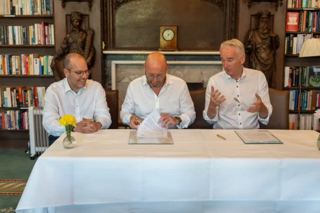 Three men sit at a table with documents to sign