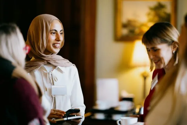 A participant wearing a headscarf speaks with a small group of people at a Wilton Park event, holding a phone and notebook while others stand nearby with coffee cups in a warmly lit indoor setting.