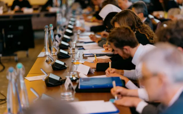 Participants take notes around the conference table during a Wilton Park dialogue, with microphones, name cards and documents visible.