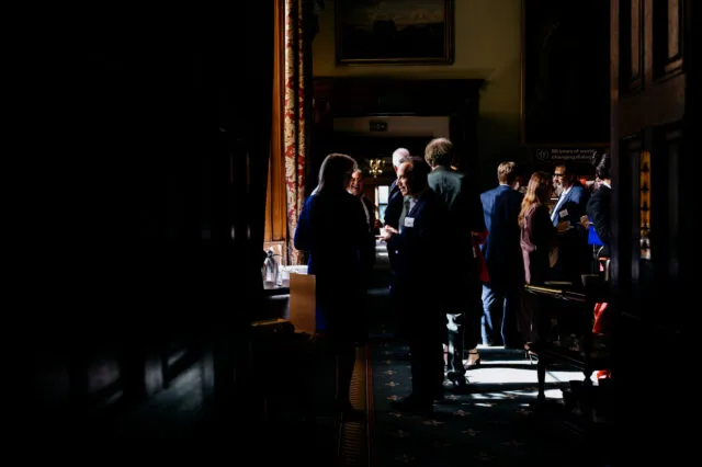 Participants are seen through a doorway standing and chatting. There are strong shadows and contrast.