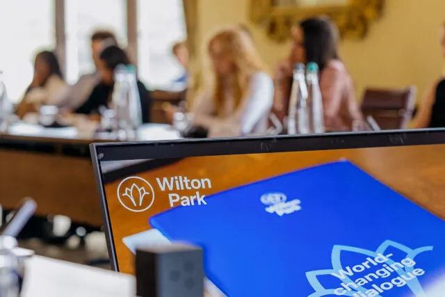 Computer monitor displaying the Wilton Park logo and a blue conference folder with the words “World-changing dialogue” visible on screen. In the background, participants are seated around the conference table at Wilton Park