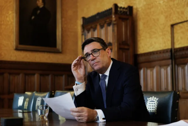 PM Andy Burnham sits at a table and holds a piece of paper.