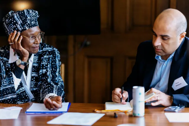 Two participants seated at a table during a breakout session.One participant, wearing patterned clothing, a matching headwrap and glasses, listens while resting a hand against the side of the face. Another participant, wearing a dark jacket and name badge, writes notes in a notebook.