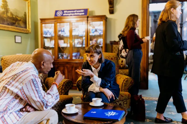 articipants networking and talking in a lounge area during a conference or workshop. Two people are seated in armchairs in the foreground engaged in conversation over coffee