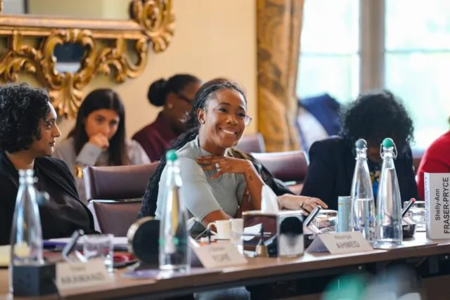 Shelly-Ann Fraser-Pryce is sat at the conference table at Wilton Park. She is smiling and has her hand raised to her chest.